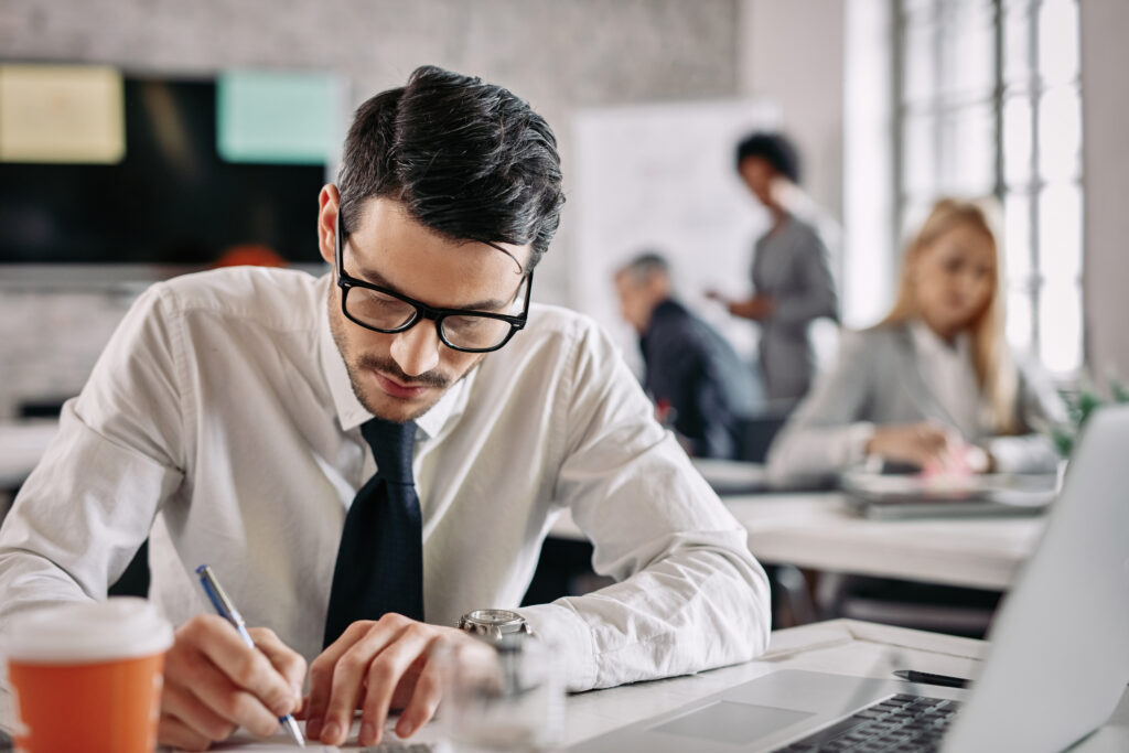 Young businessman writing at his desk while doing paperwork in the office. There are people in the background.